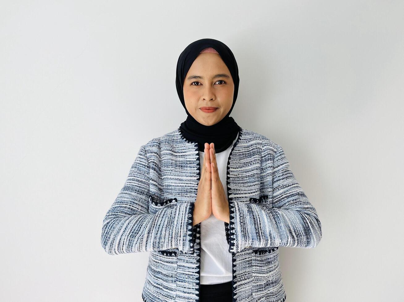 Portrait of excited Asian hijab woman in casual suit showing apologize and welcome hand gesture. Businesswoman concept. Isolated image on white background photo