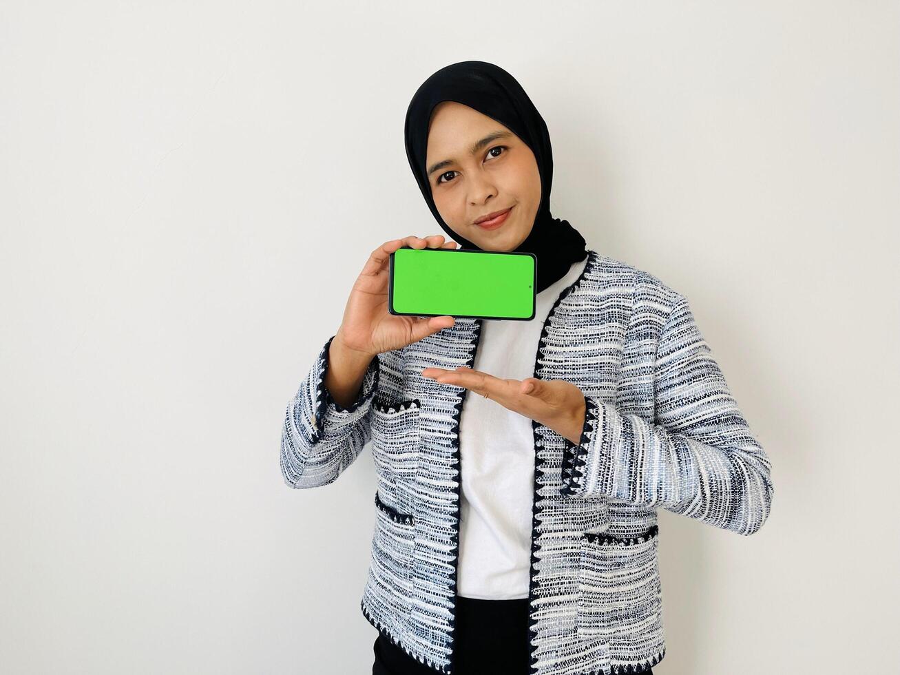 A smiling young Asian Muslim woman show smartphone green color of the screen with positive expression, dressed in tweed blazer and black hijab feeling happy isolated by white background. Mobile phone photo