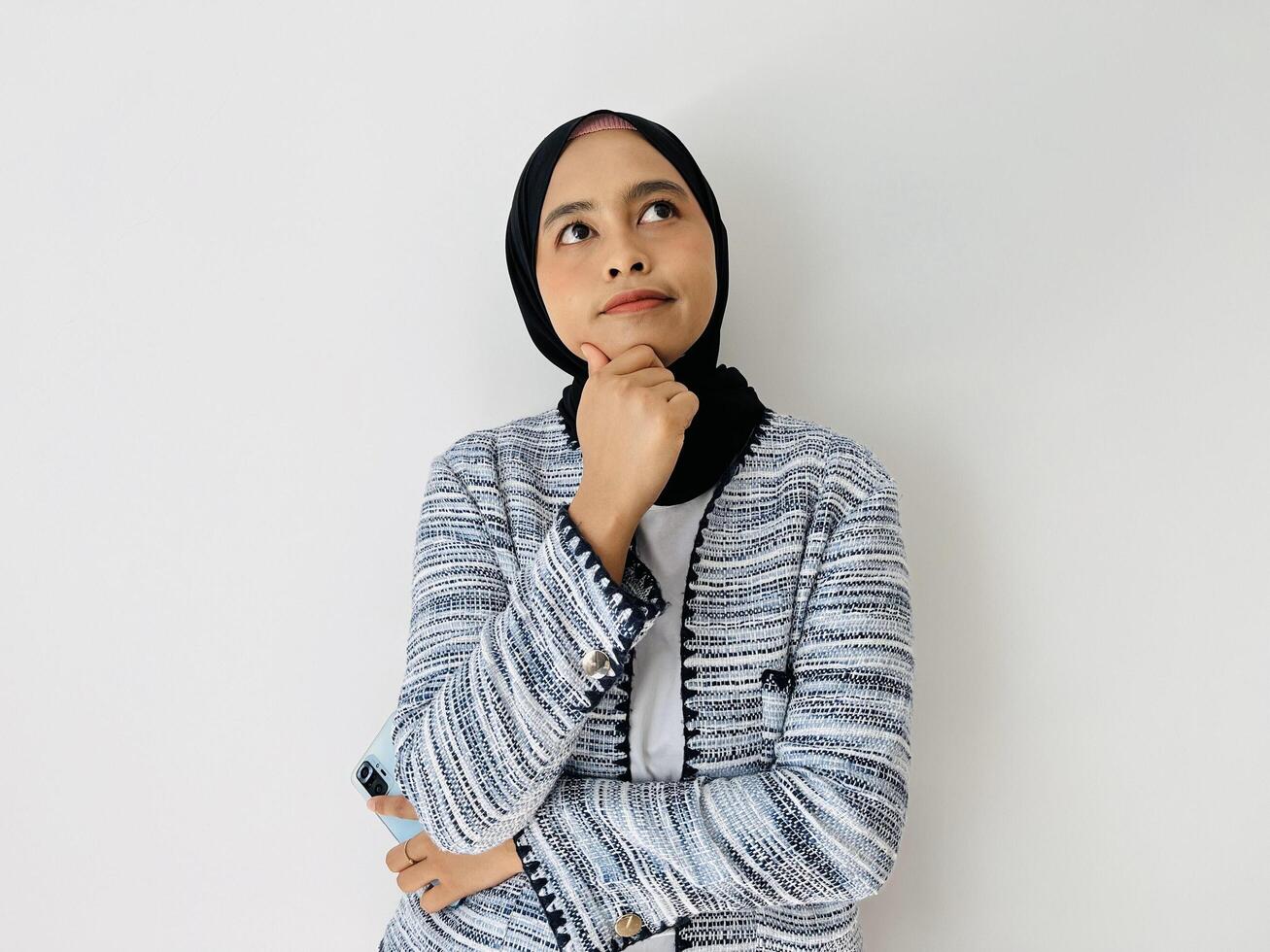 Portrait of Thoughtful Asian Muslim woman standing with her hand on chin thinking, feeling doubtful and confused, with different options, wondering which decision to make while holding phone photo