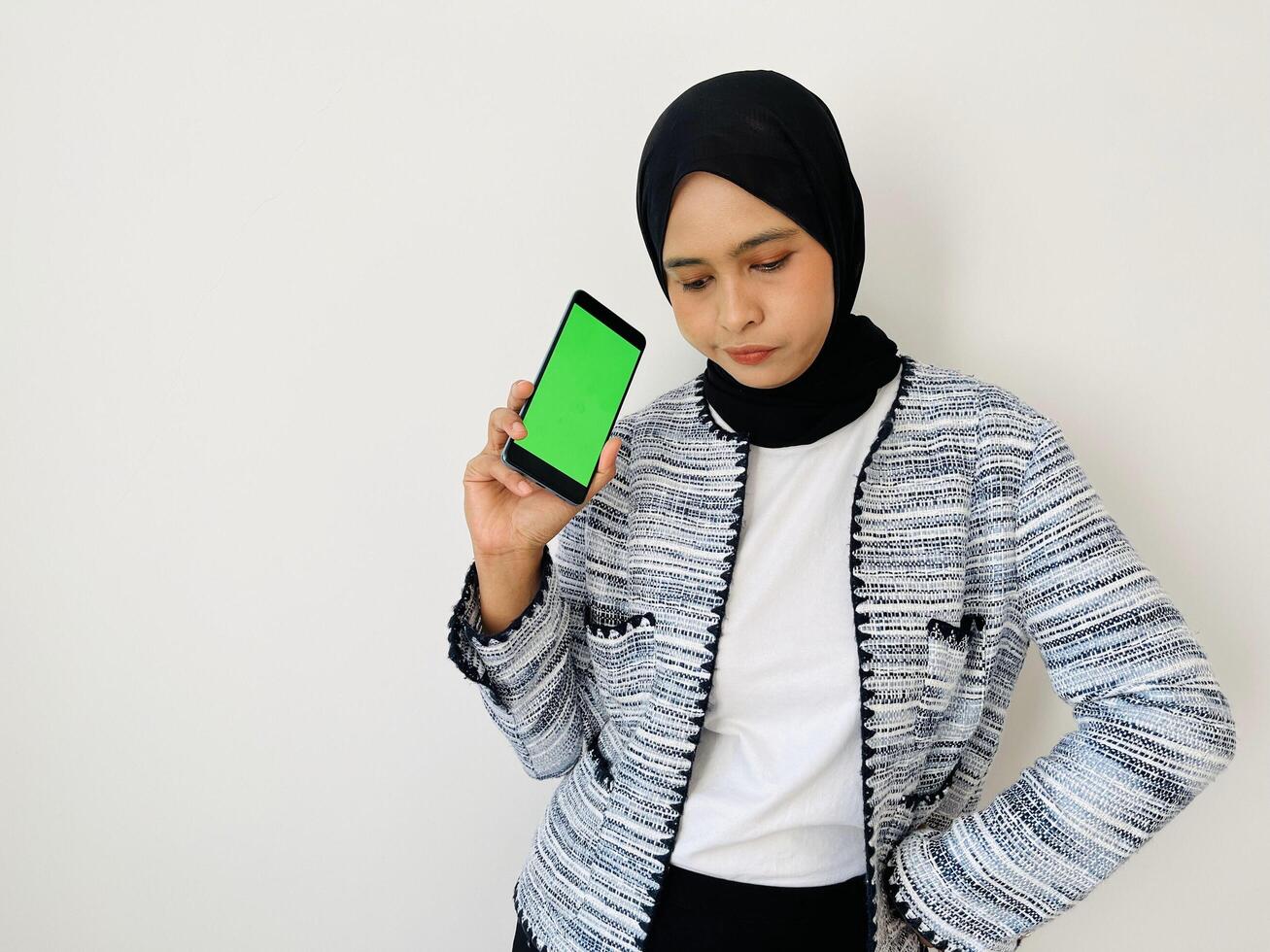 Young Asian woman holding mobile phone with green screen having doubts while looking down. Female wearing black hijab and tweed blazer isolated on white background photo