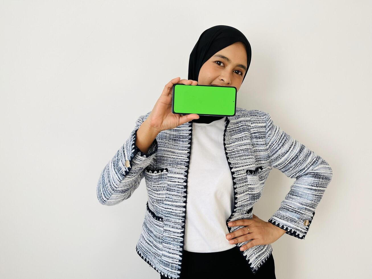 A smiling young Asian Muslim woman show smartphone green color of the screen with positive expression, dressed in tweed blazer and black hijab feeling happy isolated by white background. Mobile phone photo