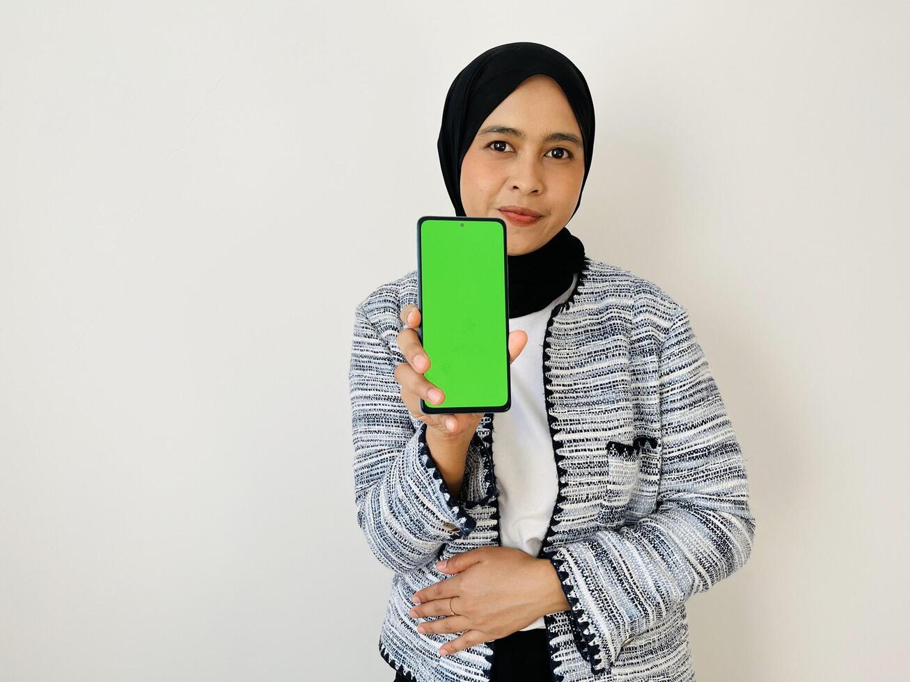 A smiling young Asian Muslim woman show smartphone green color of the screen with positive expression, dressed in tweed blazer and black hijab feeling happy isolated by white background. Mobile phone photo
