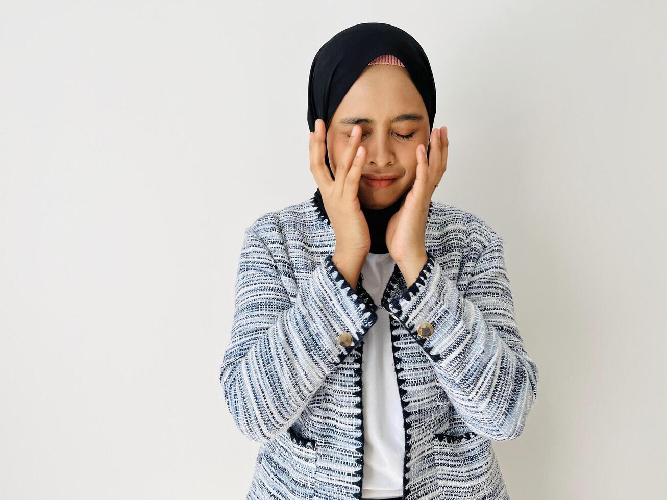 Young Asian woman in bad mood covering her face with both hands bored and feel sad. Female portrait wearing black hijab and tweed blazer isolated by white background photo
