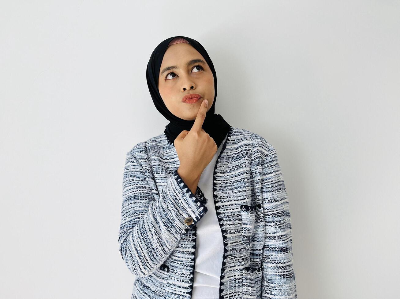 Portrait of Thoughtful Asian Muslim woman standing with her hand near lips thinking, feeling doubtful and confused, with different options, wondering which decision to make while photo