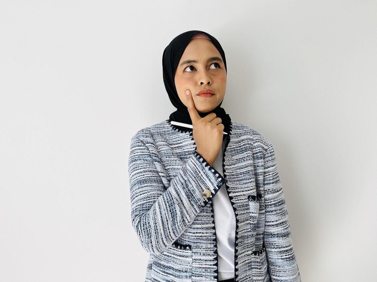 A young Asian woman wearing black hijab and tweed blazer thinks deeply about something, and keeps index finger near lips isolated by white background photo