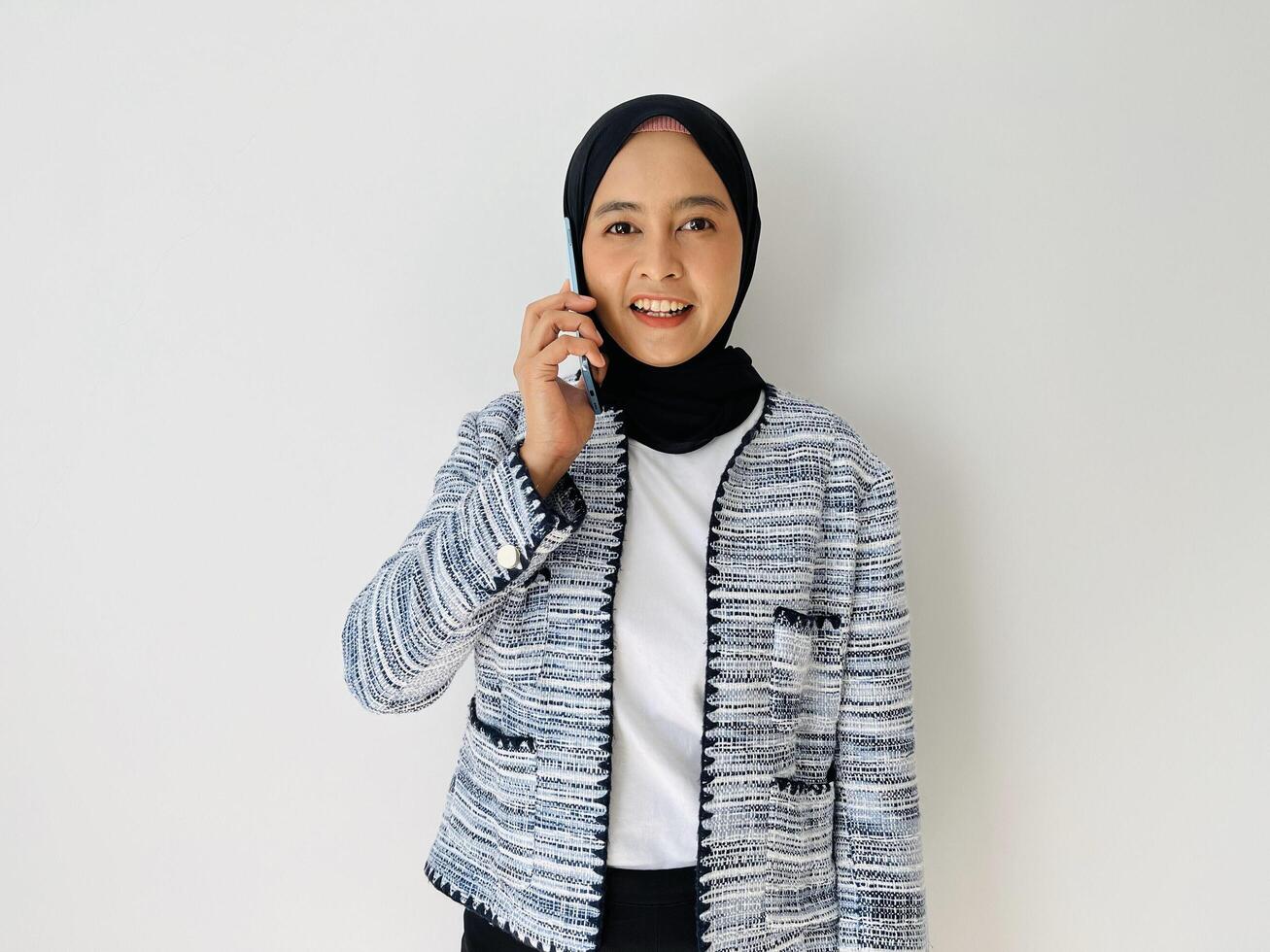 Young Asian woman smiling while using mobile phone having call. Standing female wearing black hijab and tweed blazer isolated on white background. photo