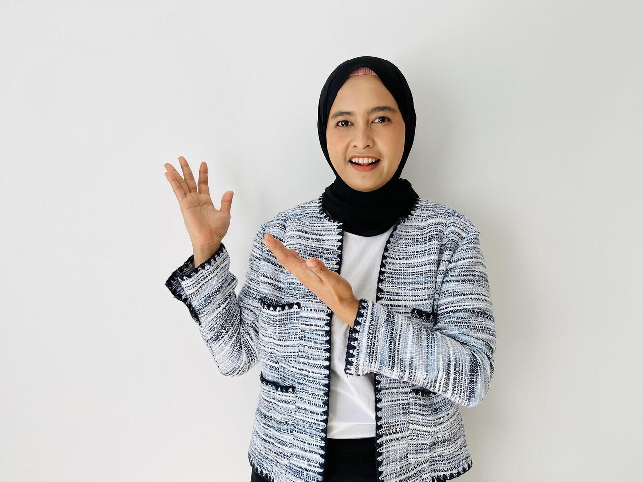 Smiling young Asian woman wearing head wearing black hijab and tweed blazer showing and pointing something standing over white background. Copy space for your text photo