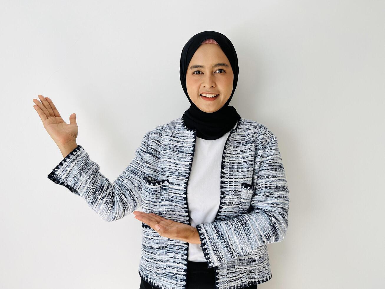 Smiling young Asian woman wearing head wearing black hijab and tweed blazer showing and pointing something standing over white background. Copy space for your text photo