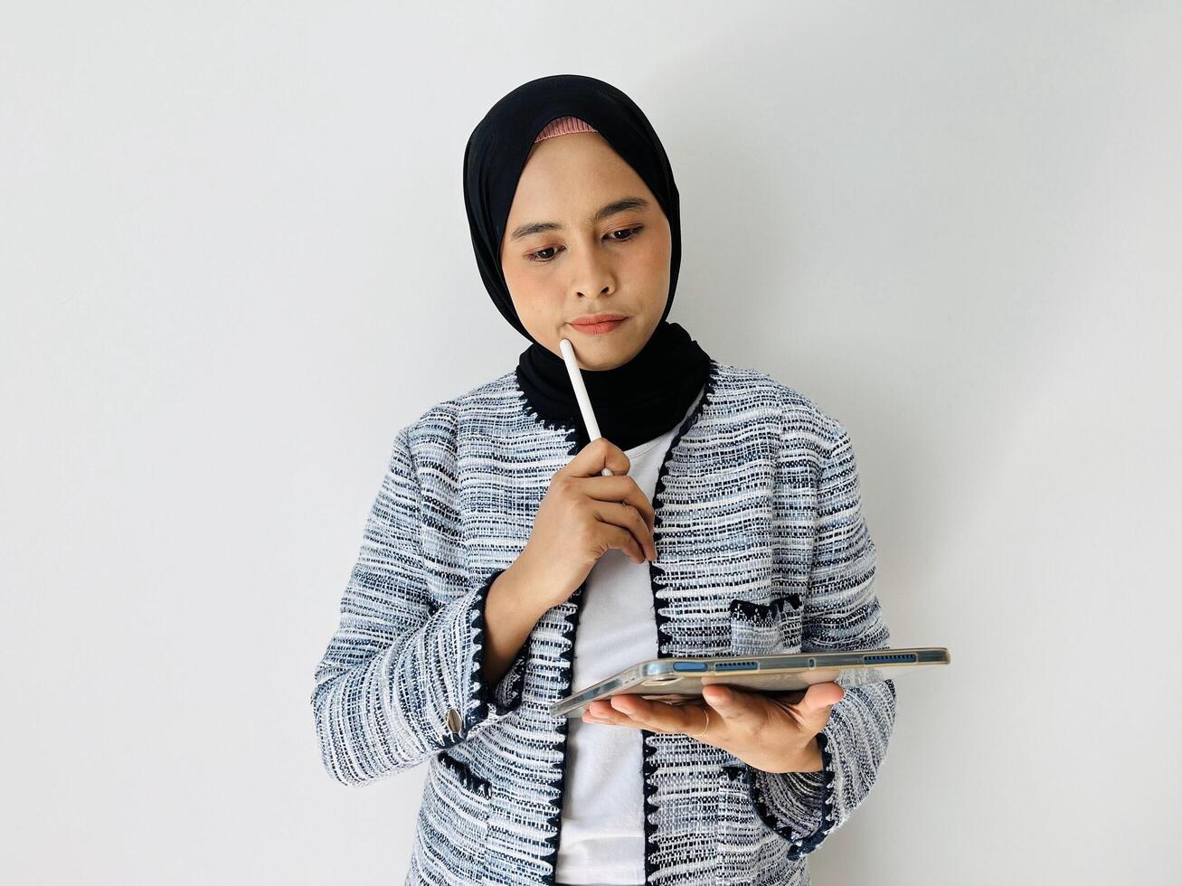 A young Asian woman wearing black hijab and tweed blazer thinks deeply about something, holding digital tablet computer and keeps the pen near lips isolated by white background photo