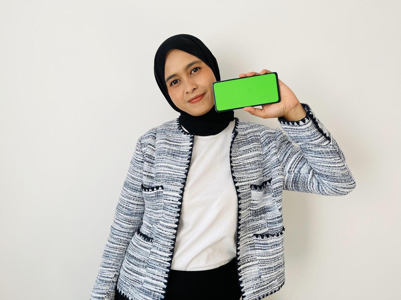 A smiling young Asian Muslim woman show smartphone green color of the screen with positive expression, dressed in tweed blazer and black hijab feeling happy isolated by white background. Mobile phone photo