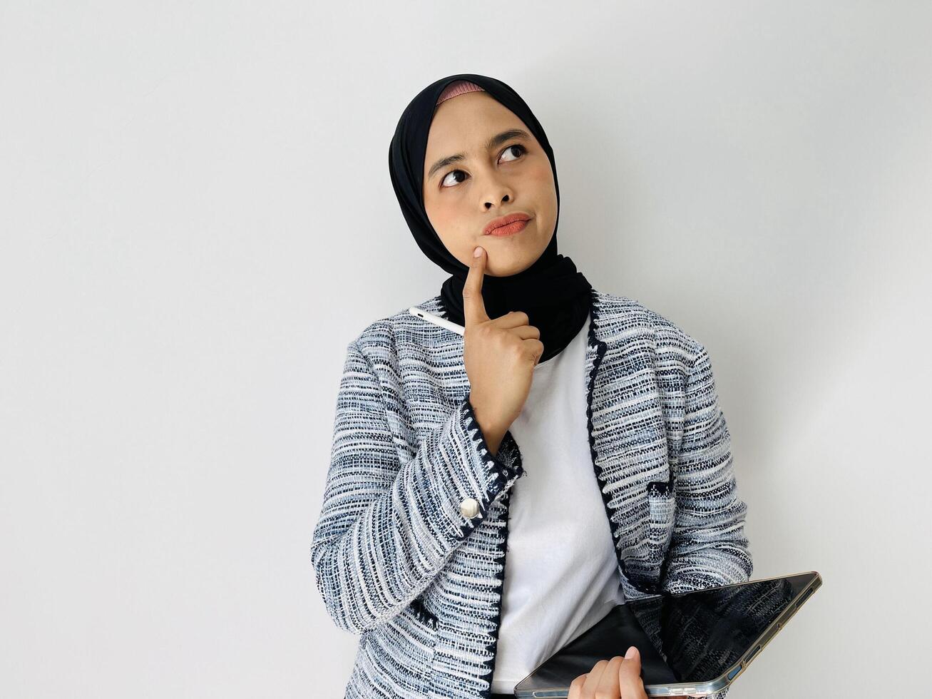 A young Asian woman wearing black hijab and tweed blazer thinks deeply about something, holding digital tablet computer and keeps the pen near lips isolated by white background photo