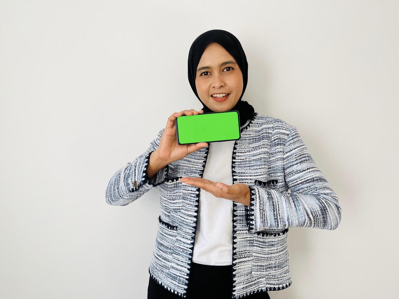 A smiling young Asian Muslim woman show smartphone green color of the screen with positive expression, dressed in tweed blazer and black hijab feeling happy isolated by white background. Mobile phone photo