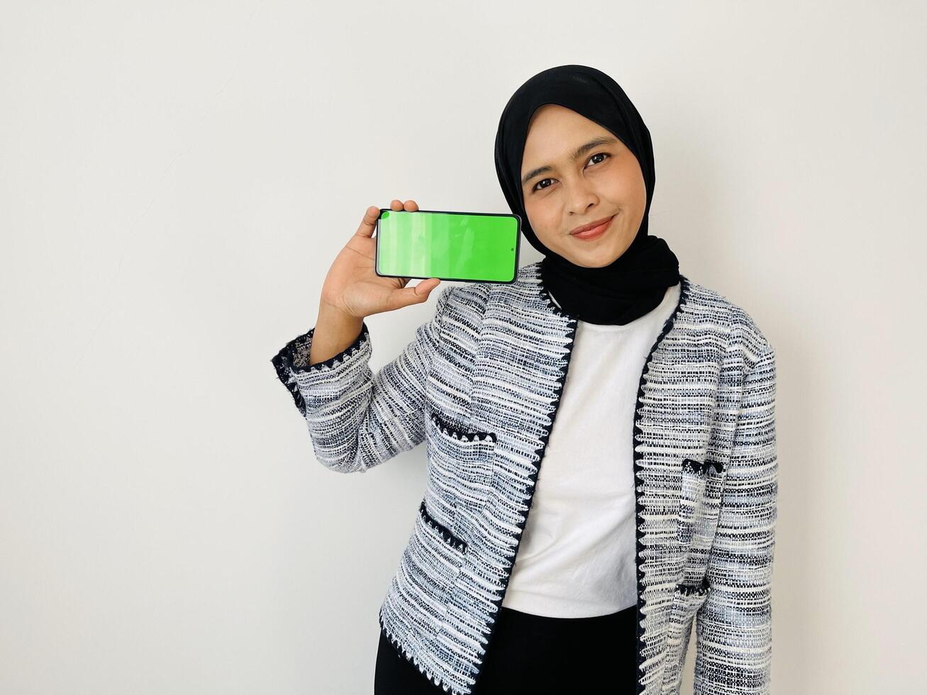 A smiling young Asian Muslim woman show smartphone green color of the screen with positive expression, dressed in tweed blazer and black hijab feeling happy isolated by white background. Mobile phone photo