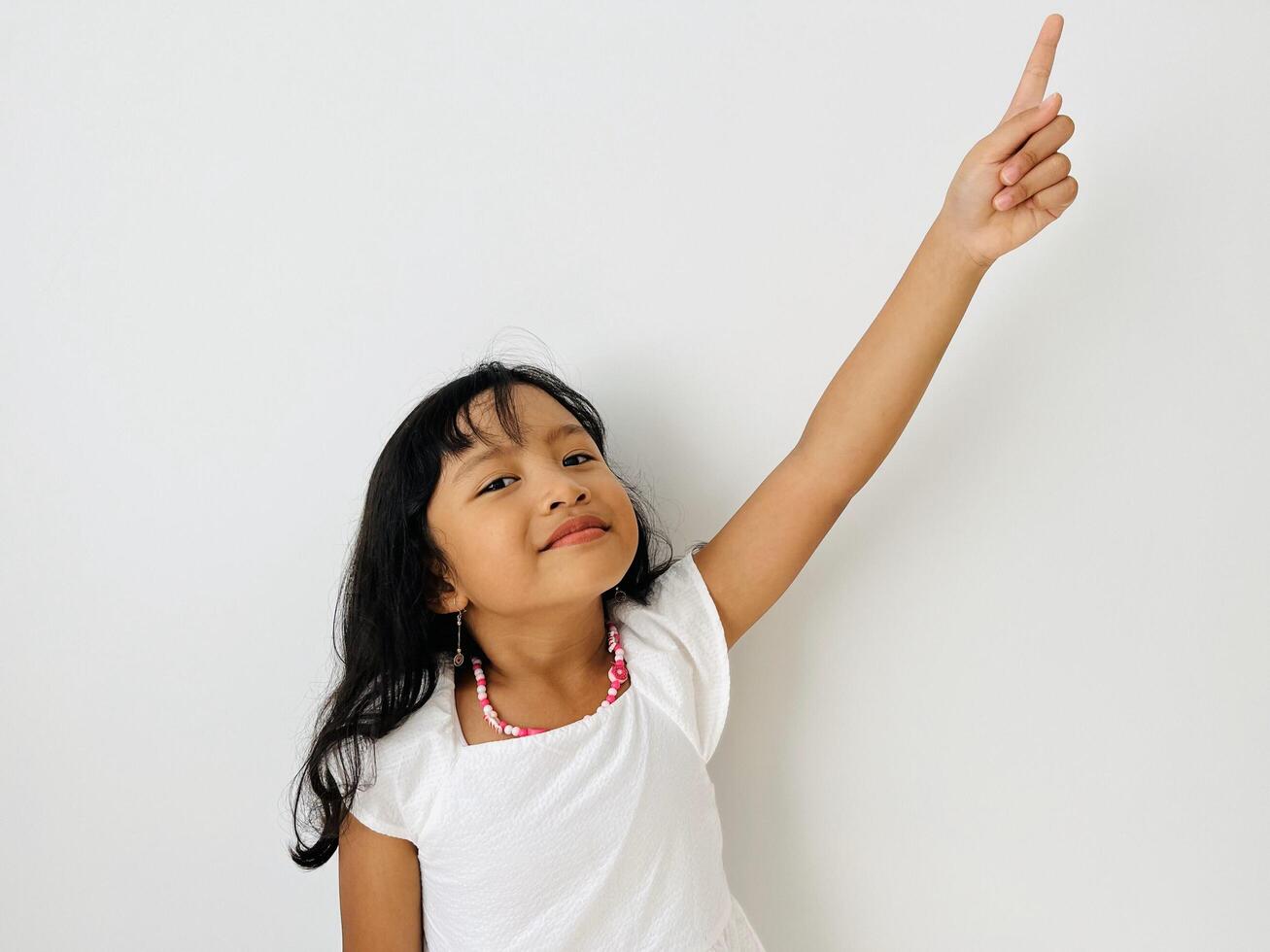 The little Asian girl raises her hand, points up with her index finger and smiles. A child in white dress on a white isolated background photo
