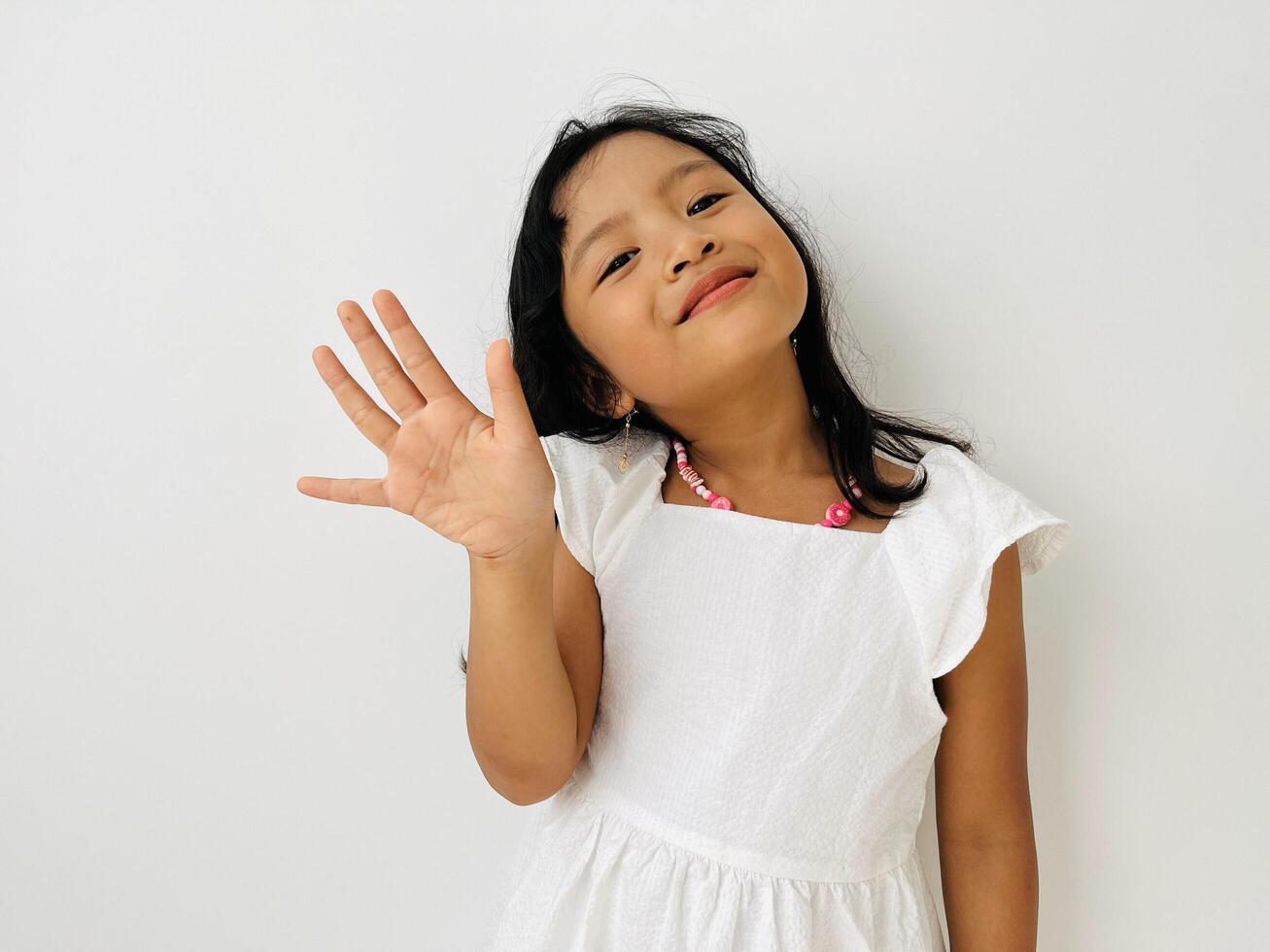 Happy cute Asian little vlogger waving hand saying hello hi looking at camera talking to webcam, smiling kid child girl making online call recording photo