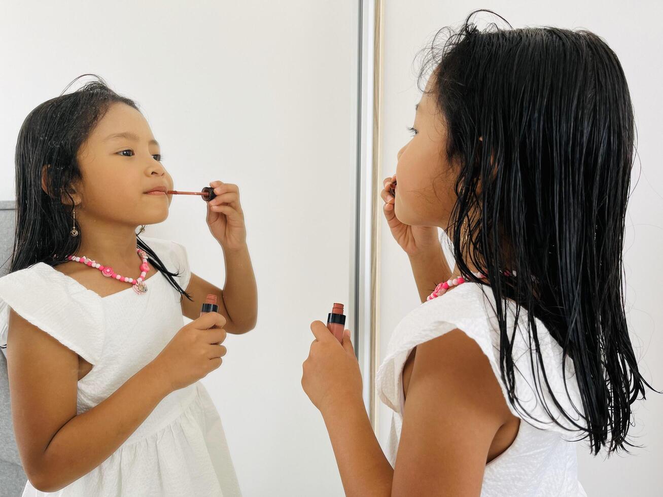 Beautiful little Asian girl in the mirror preening. Applying lipstick. A five year old in beautiful white dresses standing in front of the mirror photo