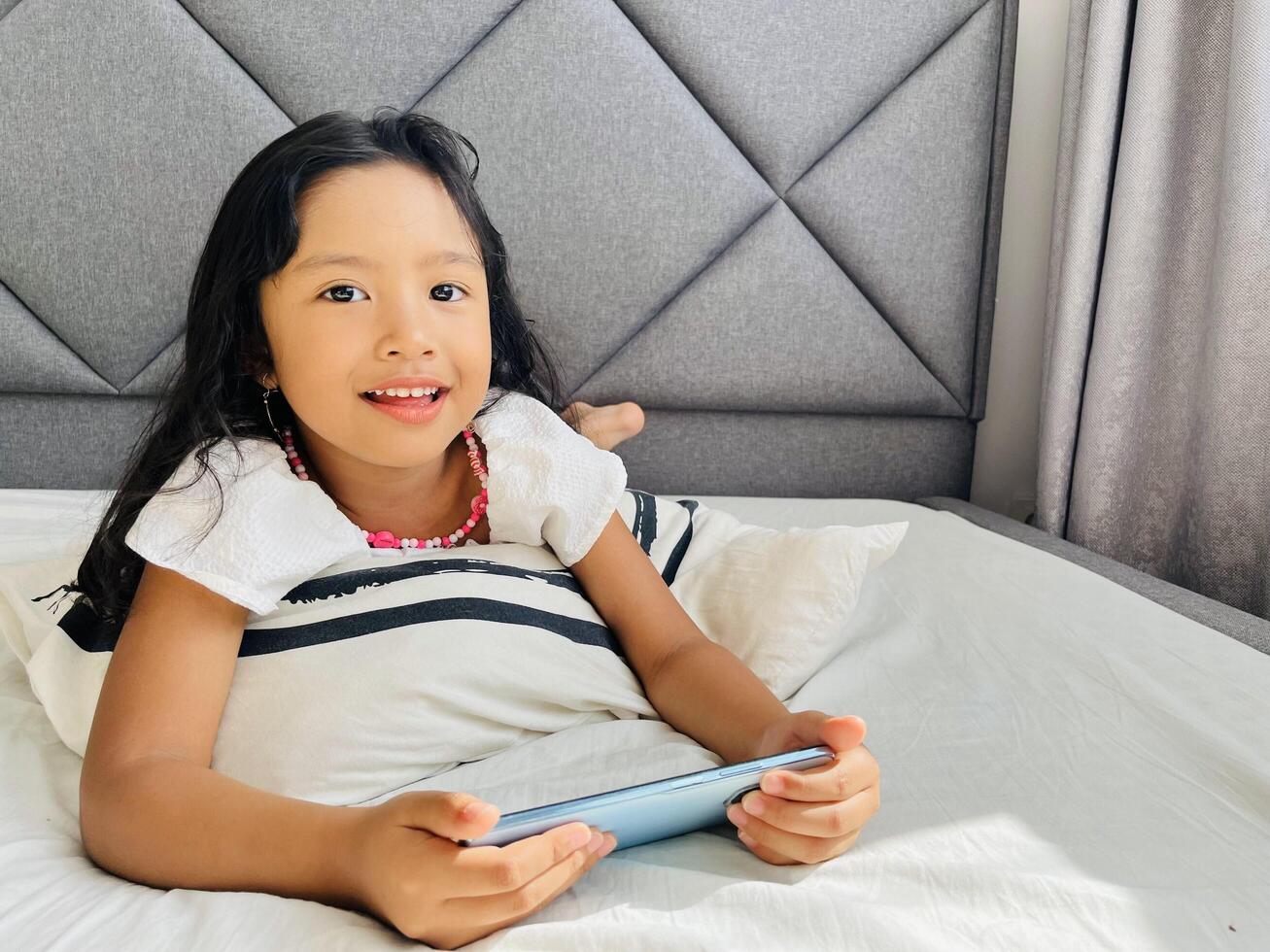 Happy adorable toddler girl playing online learning game on mobile phone. Watching cartoons on mobile phone. Asian little girl watching phone laying on the bed at home. Addicted mobile phone. photo