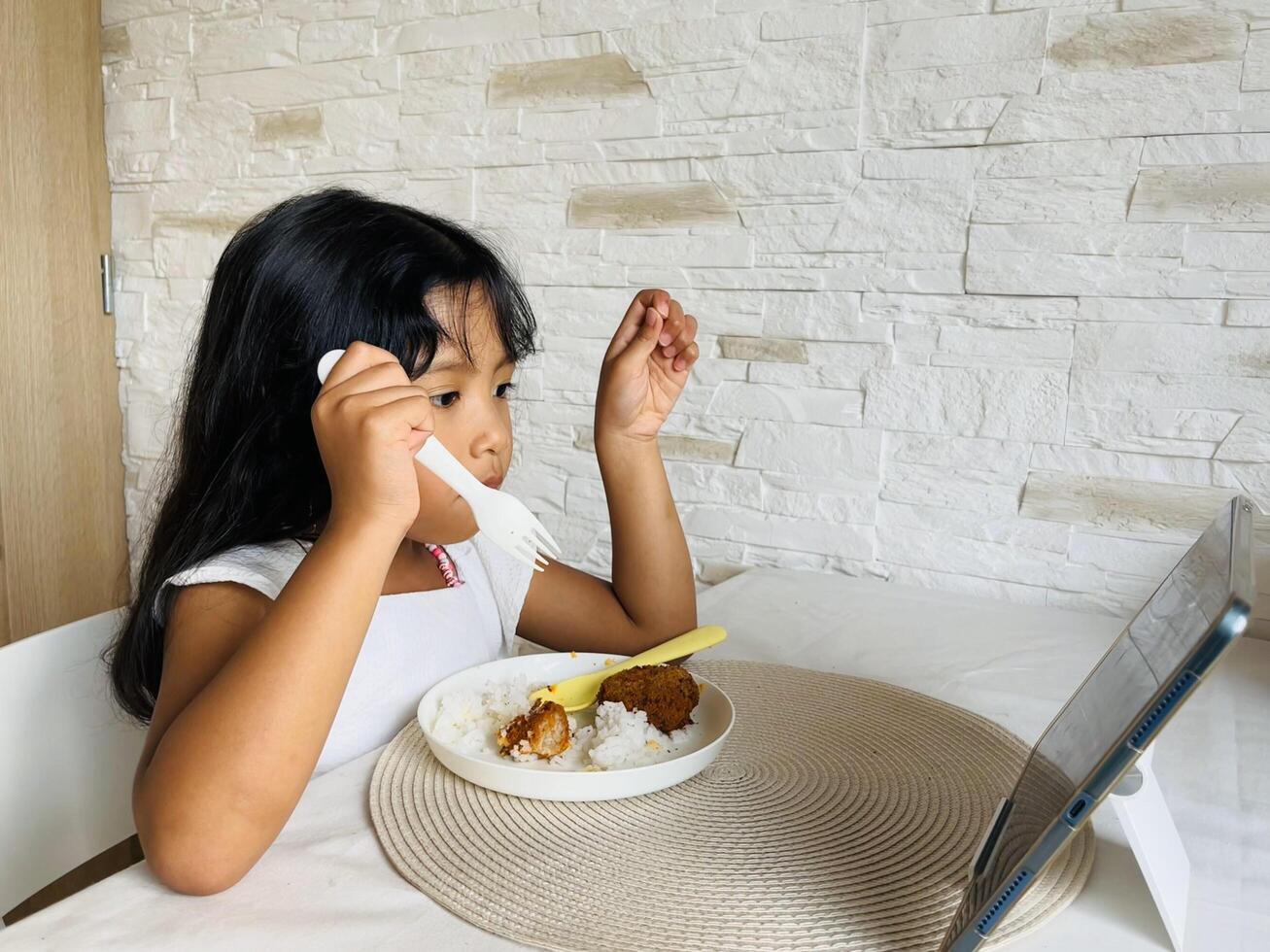 Five year old Asian girl watching cartoons on tablet while eating. Asian little girl watching tablet when dinner at home. Addicted to gadget. photo