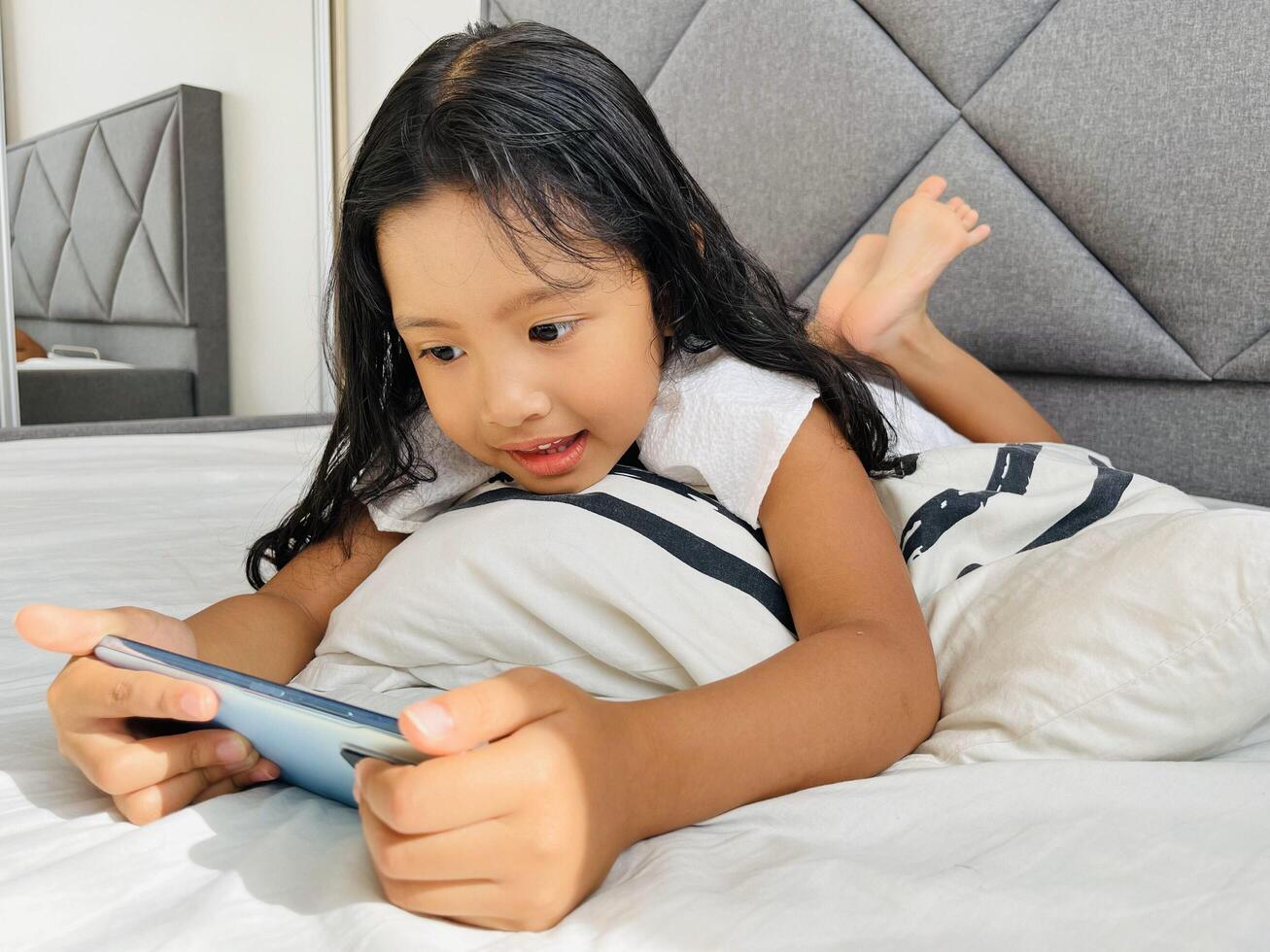 Happy adorable toddler girl playing online learning game on mobile phone. Watching cartoons on mobile phone. Asian little girl watching phone laying on the bed at home. Addicted mobile phone. photo
