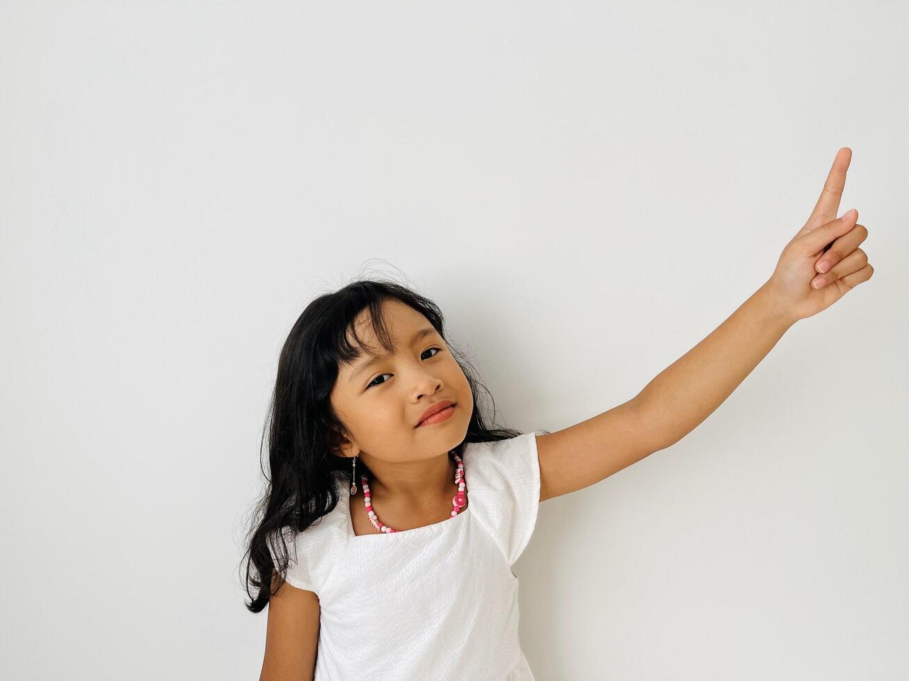The little Asian girl raises her hand, points up with her index finger and smiles. A child in white dress on a white isolated background photo