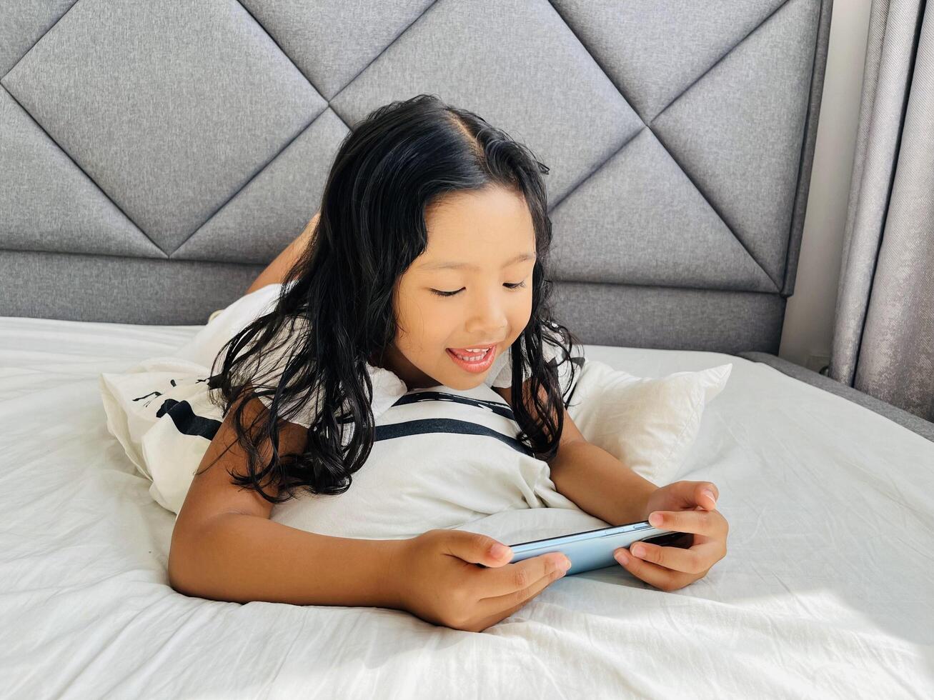 Little child girl watching cartoons on mobile phone. Asian kid watching smartphone laying on the bed at home. Little girl addicted mobile phone. Technology and internet concept photo