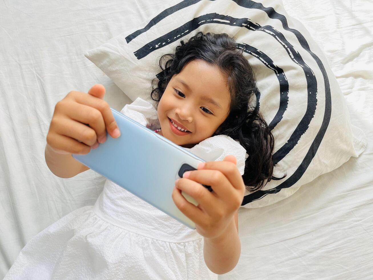 Little child girl watching cartoons on mobile phone. Asian kid watching smartphone laying on the bed at home. Little girl addicted mobile phone. Technology and internet concept photo