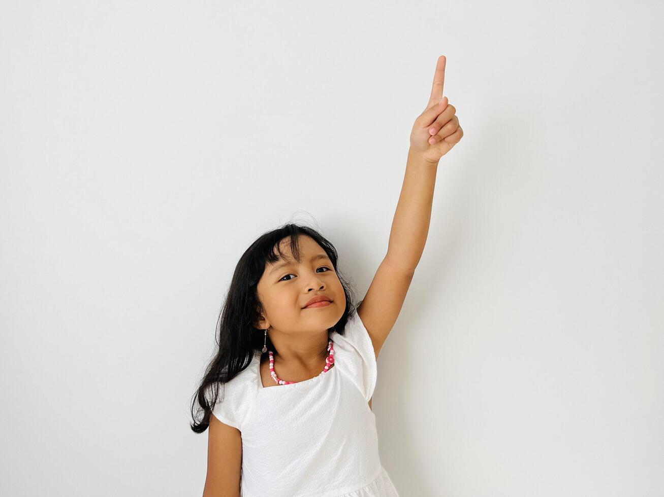 The little Asian girl raises her hand, points up with her index finger and smiles. A child in white dress on a white isolated background photo