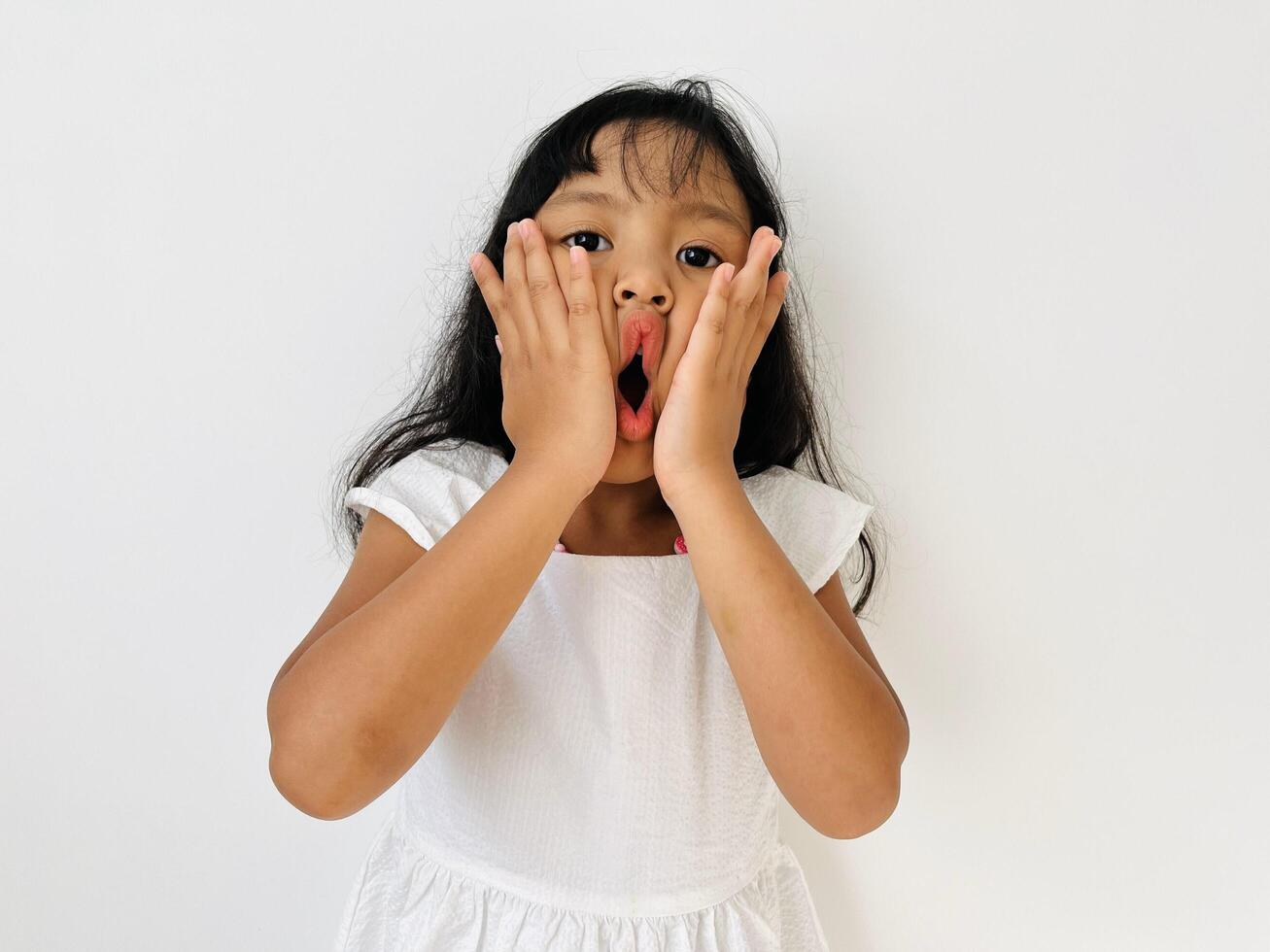 A five-year-old girl with wavy, shoulder-length black hair wearing a white dress makes a cute and silly face with her two hands pressing her lips photo