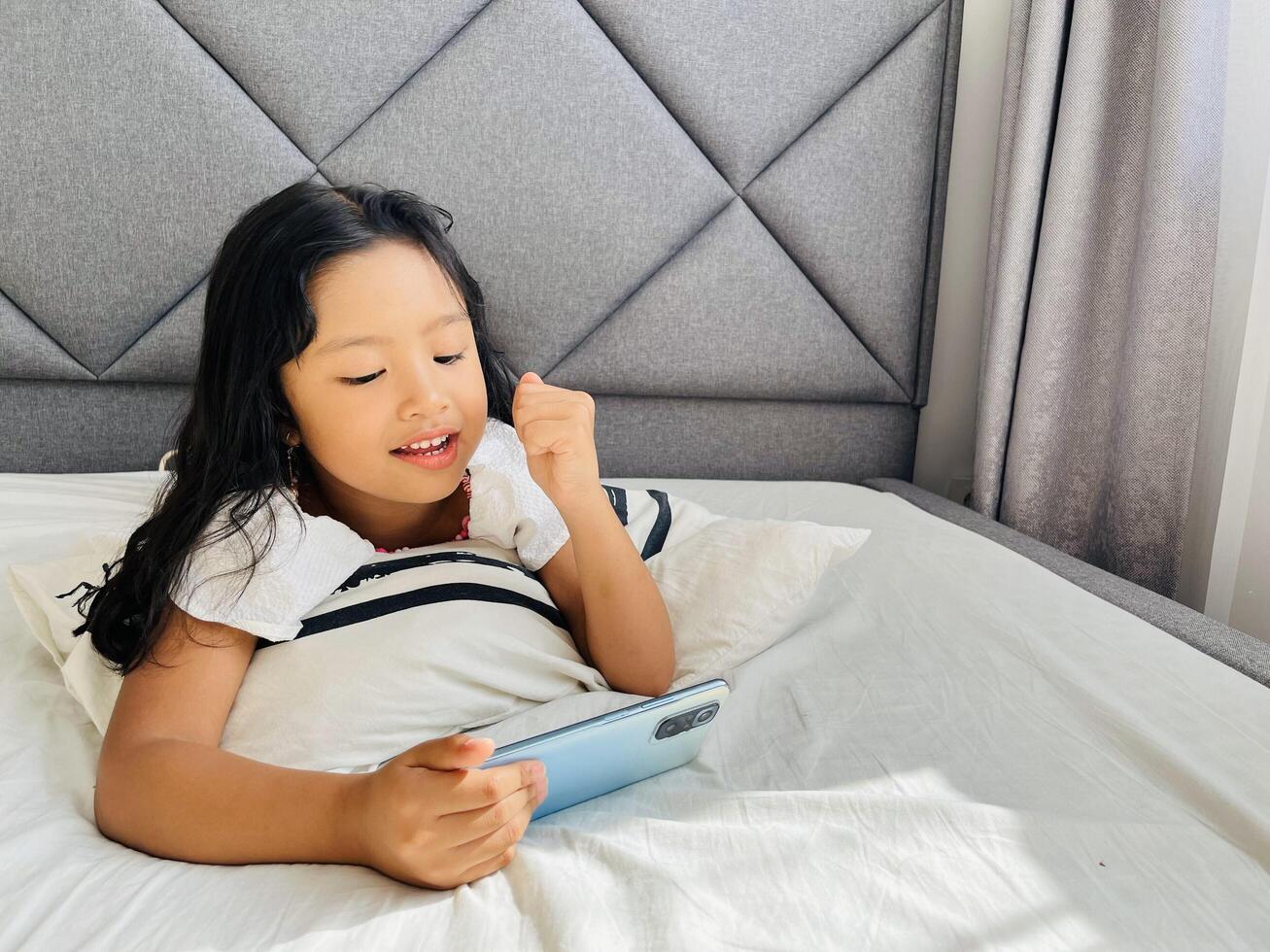 Happy adorable toddler girl playing online learning game on mobile phone. Watching cartoons on mobile phone. Asian little girl watching phone laying on the bed at home. Addicted mobile phone. photo
