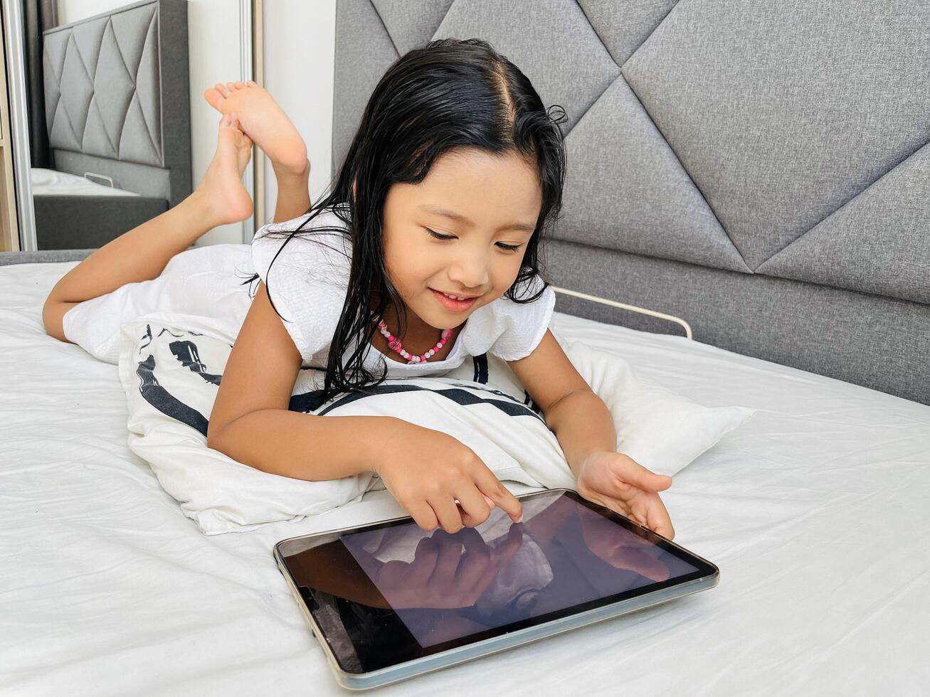 Happy adorable five year old girl playing online learning game on mobile tablet. Watching cartoons on mobile tablet. Asian little girl watching tablet laying on the bed at home. Addicted to gadget. photo