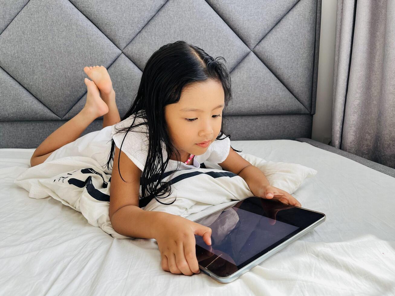 Happy adorable 5 year old girl playing online learning game on mobile tablet. Watching cartoons on mobile tablet. Asian little girl watching tablet laying on the bed at home. Addicted to gadget. photo