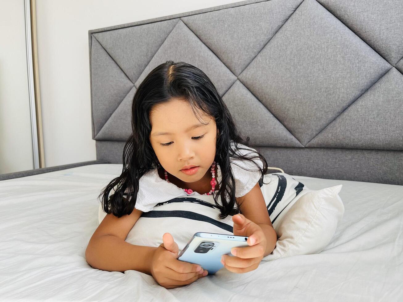 Little child girl watching cartoons on mobile phone. Asian kid watching smartphone laying on the bed at home. Little girl addicted mobile phone. Technology and internet concept photo