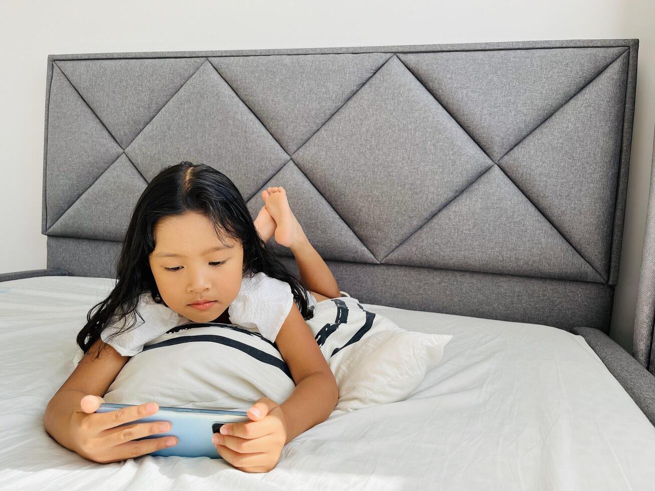 Happy adorable toddler girl playing online learning game on mobile phone. Watching cartoons on mobile phone. Asian little girl watching phone laying on the bed at home. Addicted mobile phone. photo