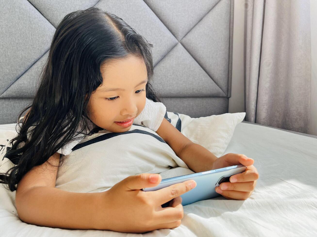 Happy adorable toddler girl playing online learning game on mobile phone. Watching cartoons on mobile phone. Asian little girl watching phone laying on the bed at home. Addicted mobile phone. photo