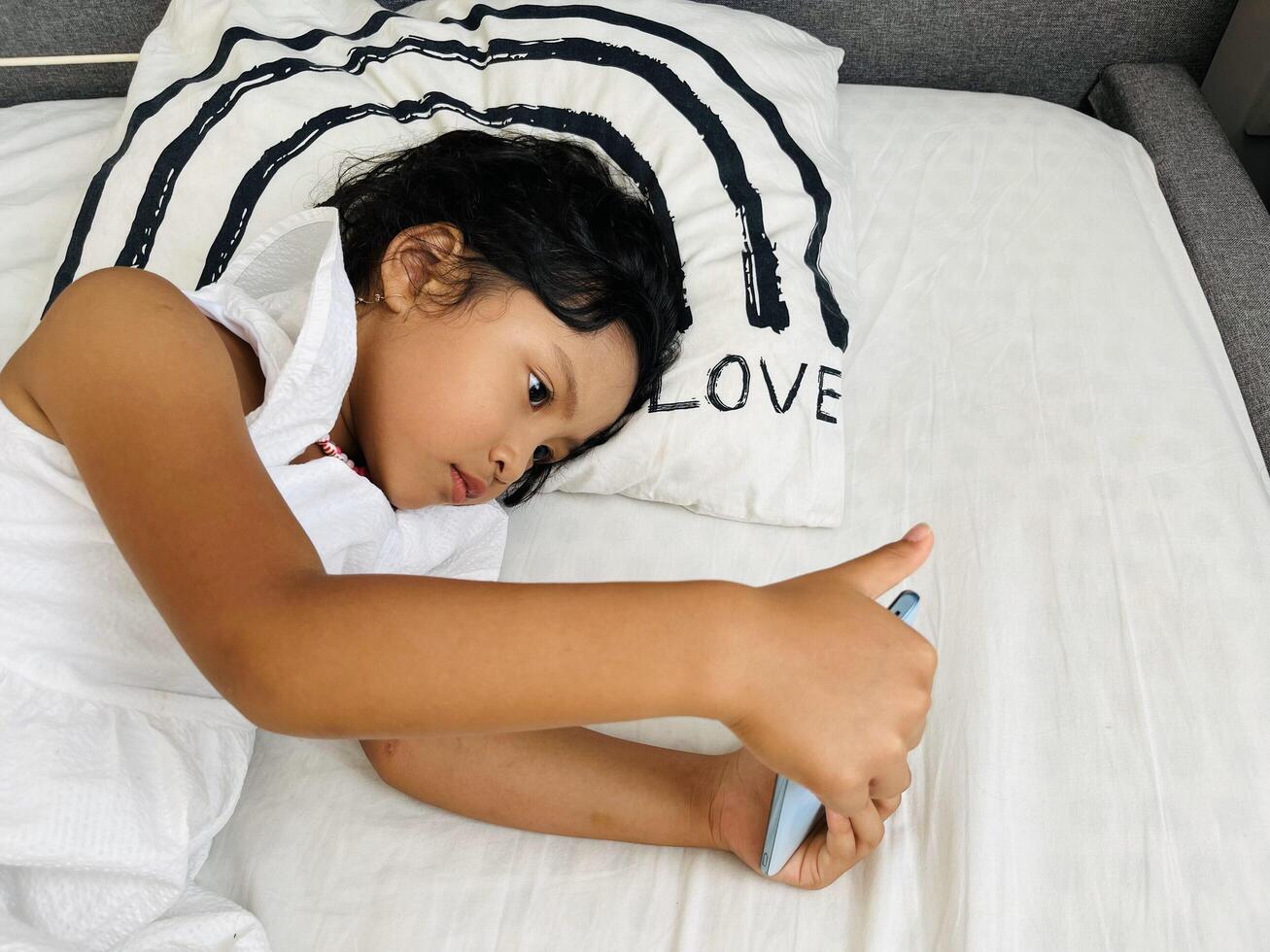 Happy adorable toddler girl playing online learning game on mobile phone. Watching cartoons on mobile phone. Asian little girl watching phone laying on the bed at home. Addicted mobile phone. photo