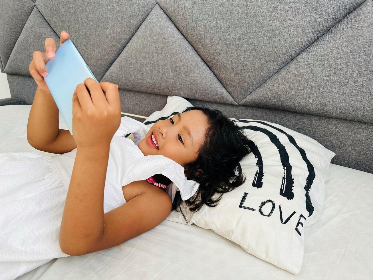 Happy adorable toddler girl playing online learning game on mobile phone. Watching cartoons on mobile phone. Asian little girl watching phone laying on the bed at home. Addicted mobile phone. photo