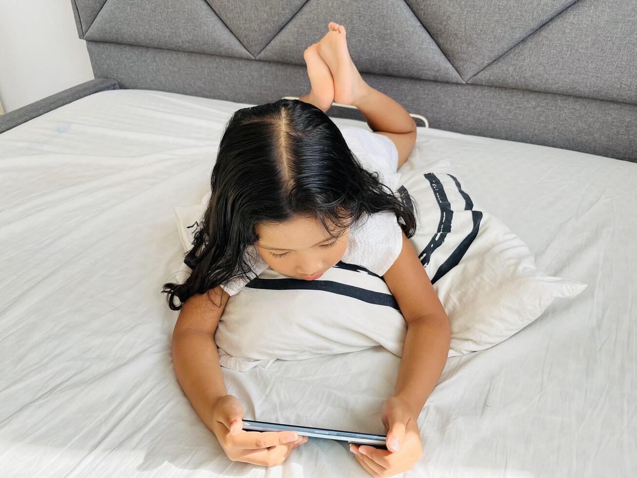 Little child girl watching cartoons on mobile phone. Asian kid watching smartphone laying on the bed at home. Little girl addicted mobile phone. Technology and internet concept photo