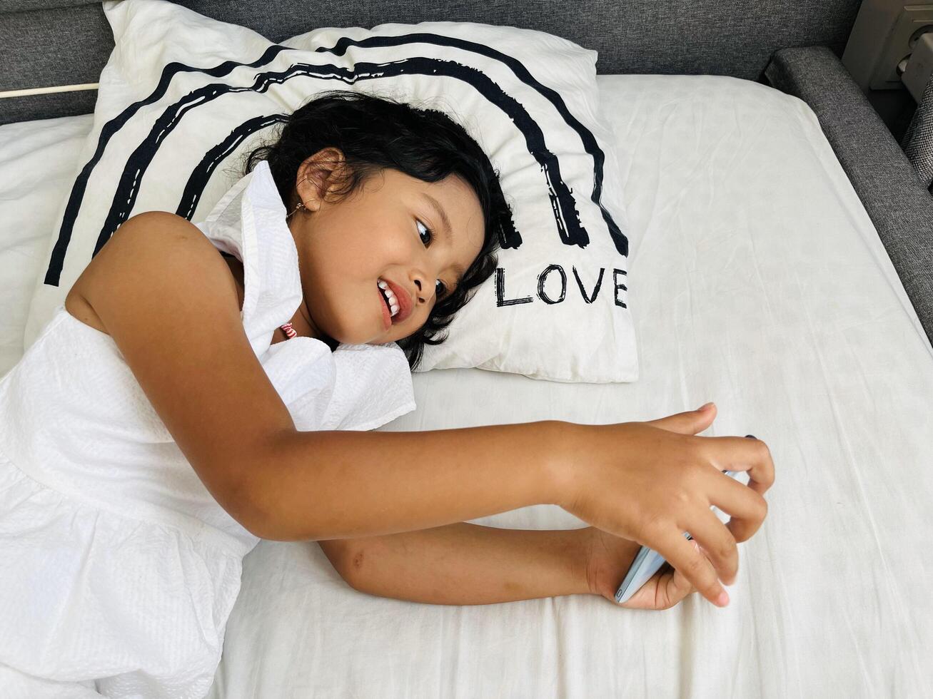 Happy adorable toddler girl playing online learning game on mobile phone. Watching cartoons on mobile phone. Asian little girl watching phone laying on the bed at home. Addicted mobile phone. photo