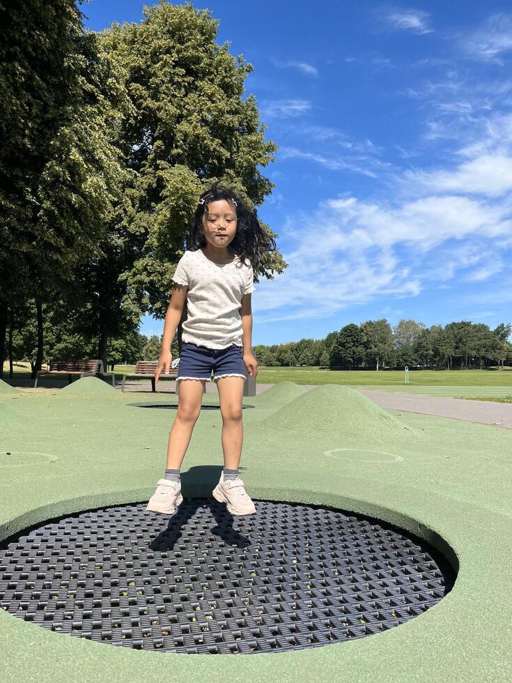 Little Asian child enjoys jumping on trampoline. Happy little girl bouncing on a trampoline on the playground in a park in the afternoon. Outdoor activity photo
