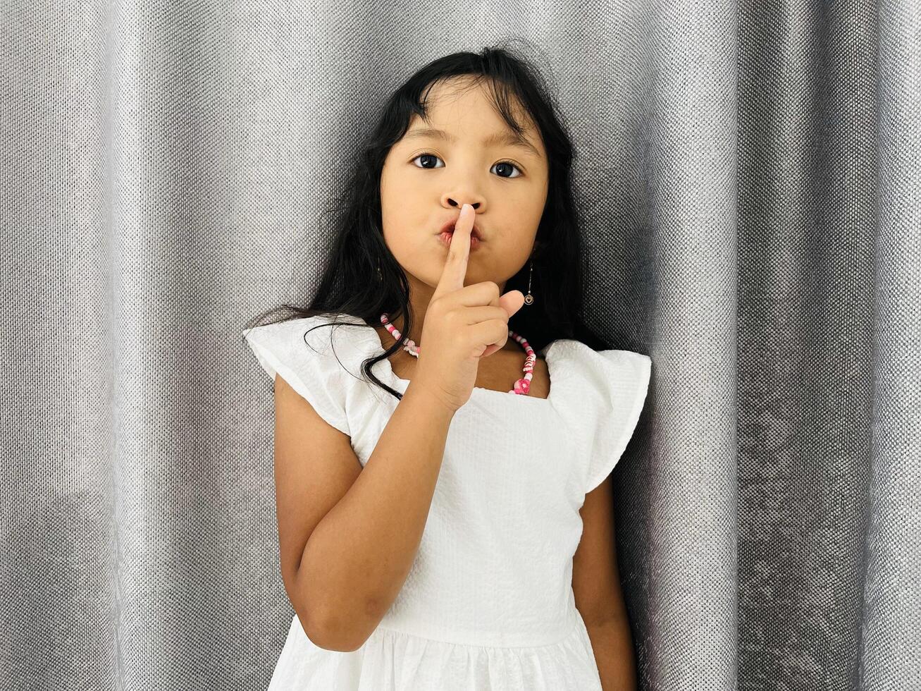 Emotional child girl has secret. Little girl with wonder face keeps finger on lips, shows silence sign, asks to be quiet and stop make loud noise. Silent gesture. photo