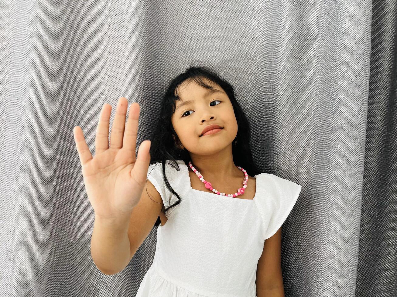 Cute adorable 5 years old girl happily shows an open hand. Little kid wearing a white dress is holding up five fingers in front of gray curtains photo