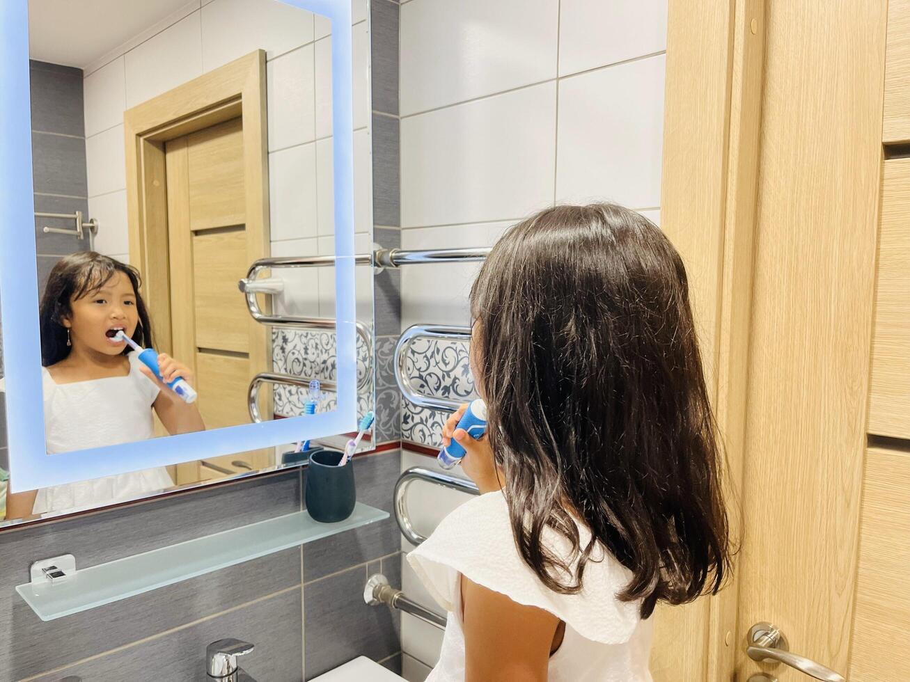 A happy little Asian girl brushes her teeth in front of a mirror in the bathroom. Morning routine. Hygiene photo
