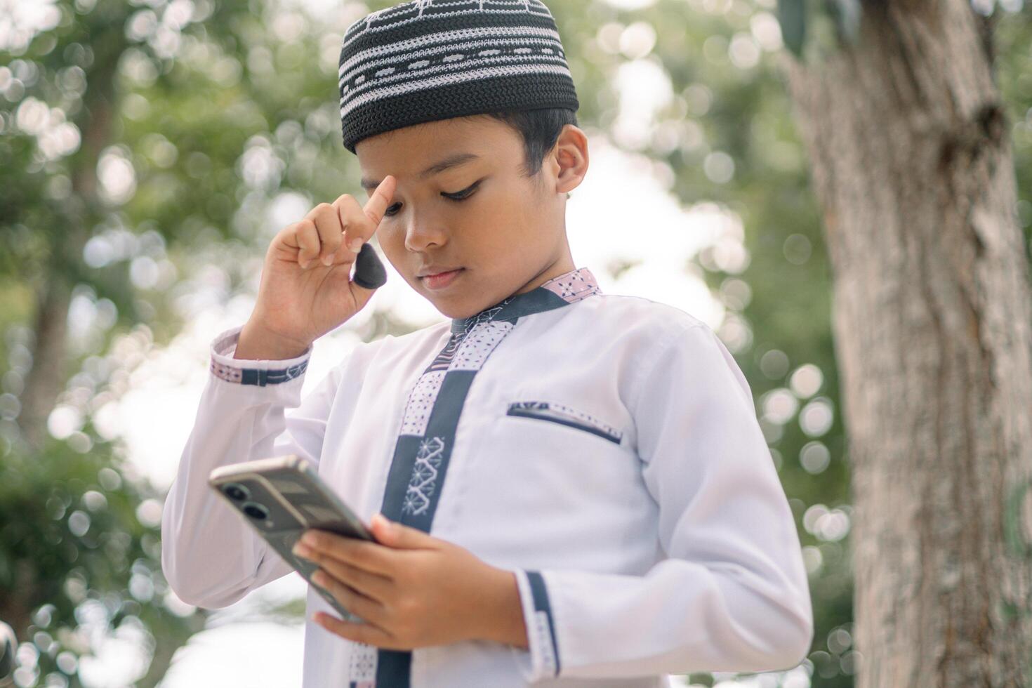 Baubau, Indonesia, June 15 2024 - Asian Muslim boy wearing a cap is playing with his smartphone photo
