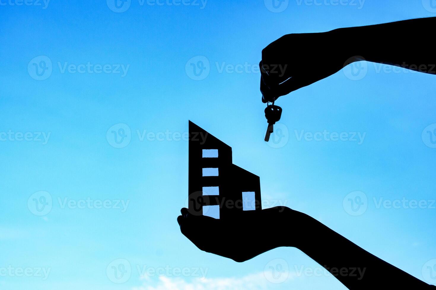 Silhouette of man hand holding small icon building model and keys against sky sunset background. hands holding symbol paper of building for concept of dream to buy house family and real estate. photo
