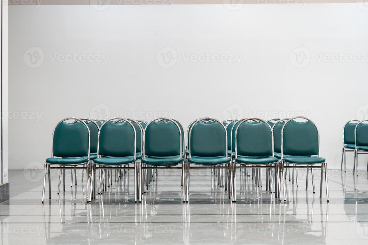 Many empty green leather chairs in a wide hall room, lights on. A floor reflect the objects. Give a feeling of emptiness. photo