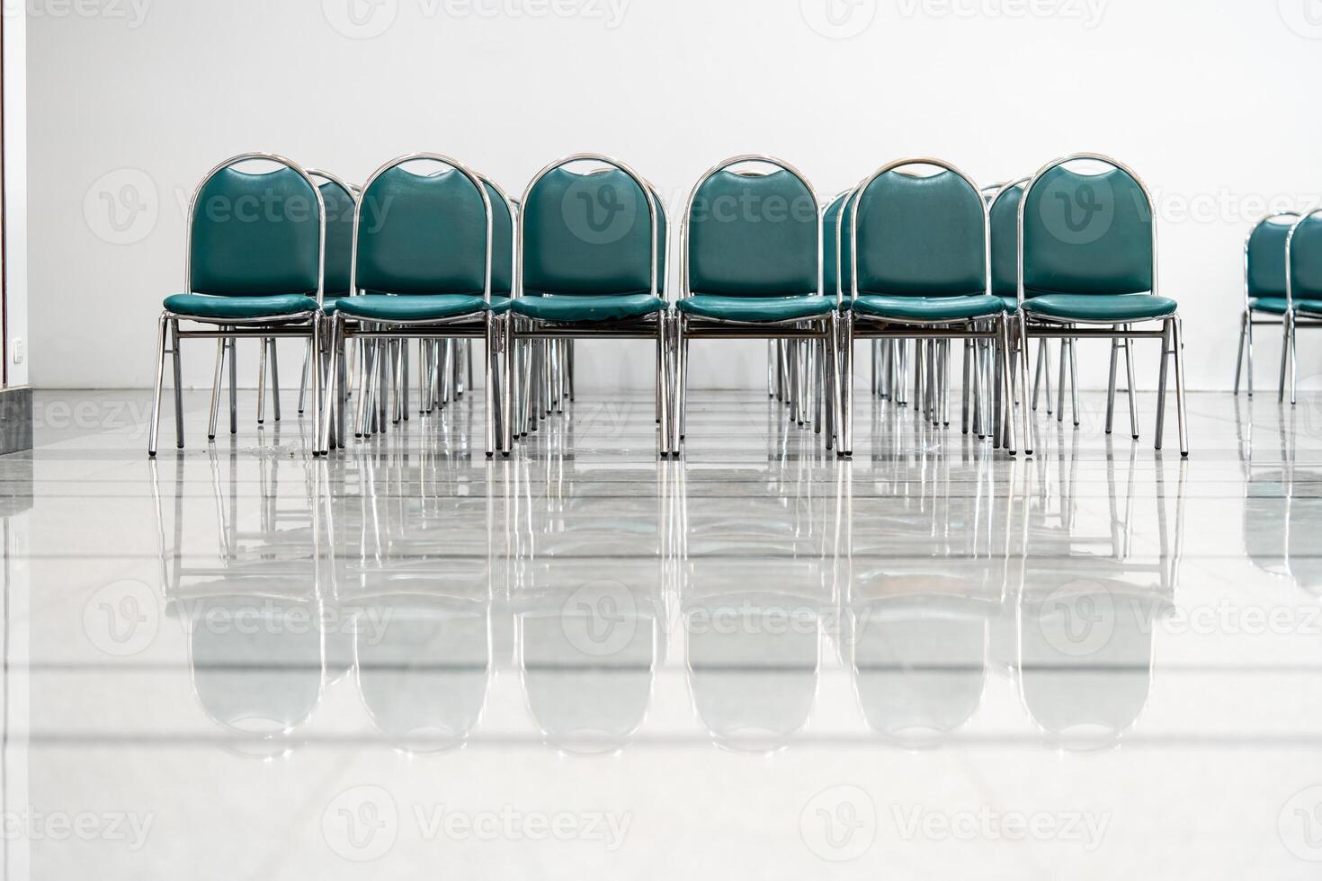 Many empty green leather chairs in a wide hall room, lights on. A floor reflect the objects. Give a feeling of emptiness. photo