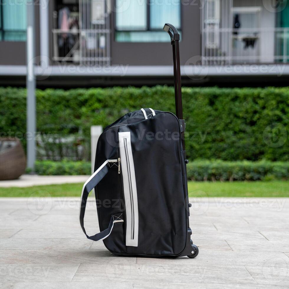 A traveler's black and white stripe suitcase with wheels was left alone on the footpath with the blurred bushes background of a luxury condo. photo