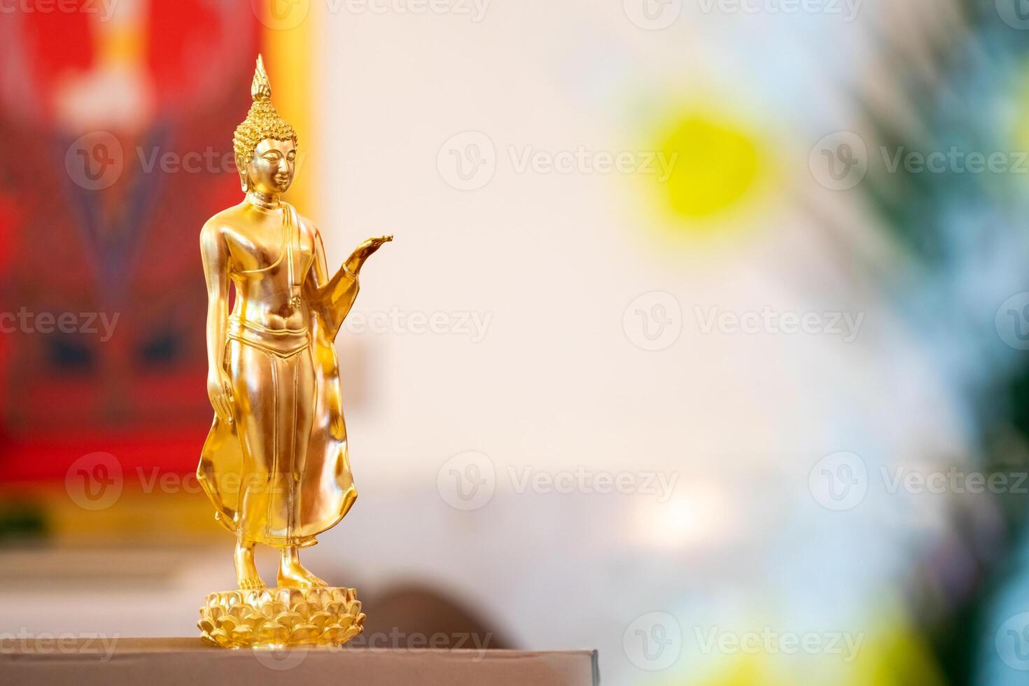 Standing Buddha statue is placed on top of a rectangular object in front of a red framed picture and tree house. photo