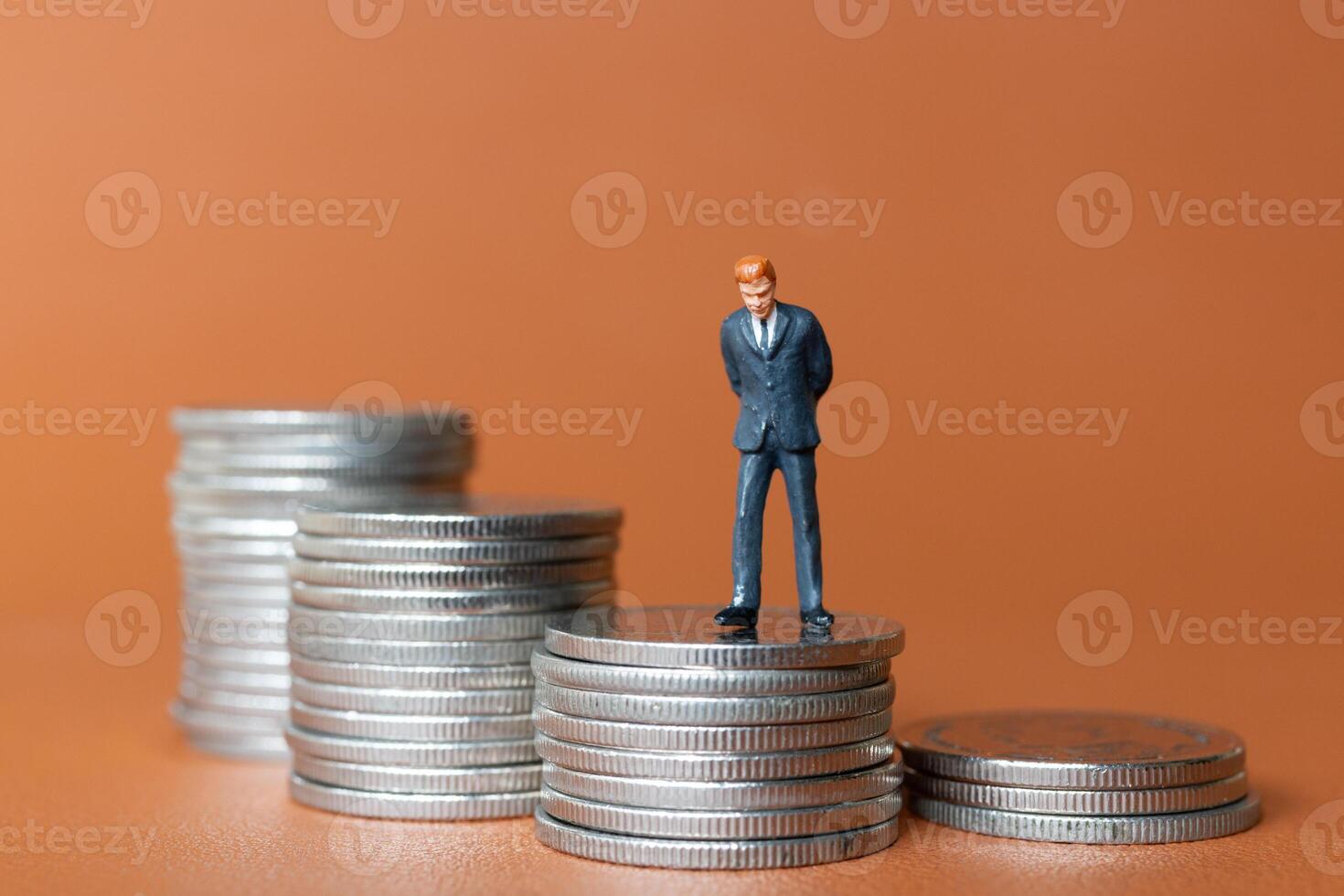 Miniature people A businessman in a suit stands with several stacks of silver coins photo