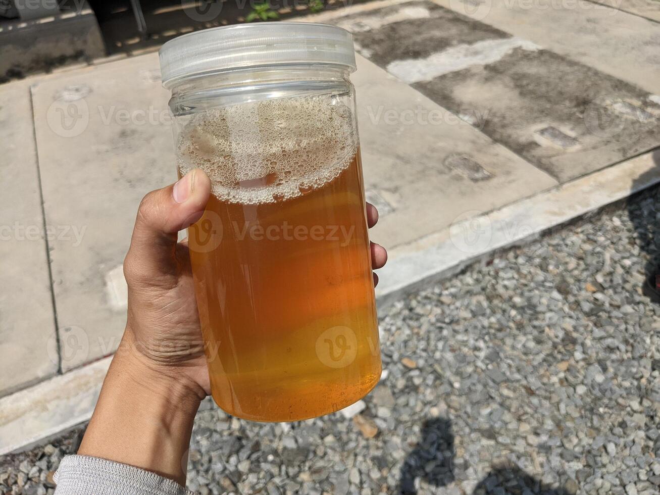 Hand holding a jar of industry oil sample. A close up of a hand holding a glass jar filled with a yellow liquid and some foam, with a man standing in the background. photo